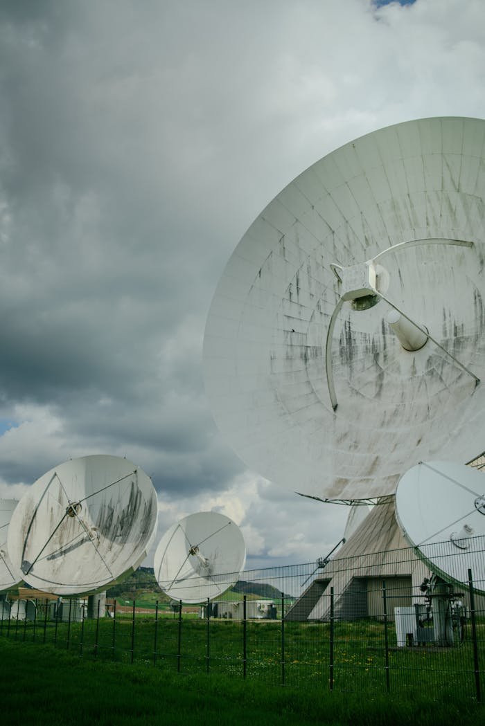 Multiple large satellite dishes at a communication facility under a cloudy sky.